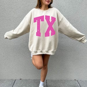May include: A beige oversized sweatshirt with the pink letters "TX" printed on the front. The letters have a distressed, vintage look. The sweatshirt has long sleeves and a crew neck.