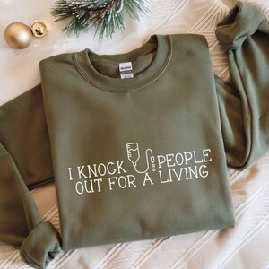 May include: Olive green sweatshirt with the text "I knock people out for a living" in white. The graphic includes an IV bag. The sweatshirt is folded on a white surface with holiday decorations.