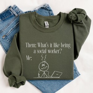 May include: An olive green sweatshirt with the text "Them: What's it like being a social worker? Me:" and a stick figure with a stressed expression, sitting at a laptop. The sweatshirt is folded on top of a pair of blue jeans.