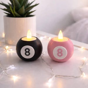 May include: Two round LED candles, resembling billiard balls, with a flickering yellow light. One is black, the other pink, both displaying the number 8. A succulent plant in a white pot is in the background. The candles are on a white surface.