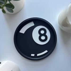 May include: A round black tray with a white eight ball design. The number eight is in white with a black outline. The tray has white accent lines. This decorative tray is suitable for various uses. The tray is a stylish item.