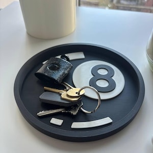 May include: A black circular tray containing keys, a black and silver lock, and a white and black number eight ball. The tray sits on a white surface, with a white plant pot in the background.