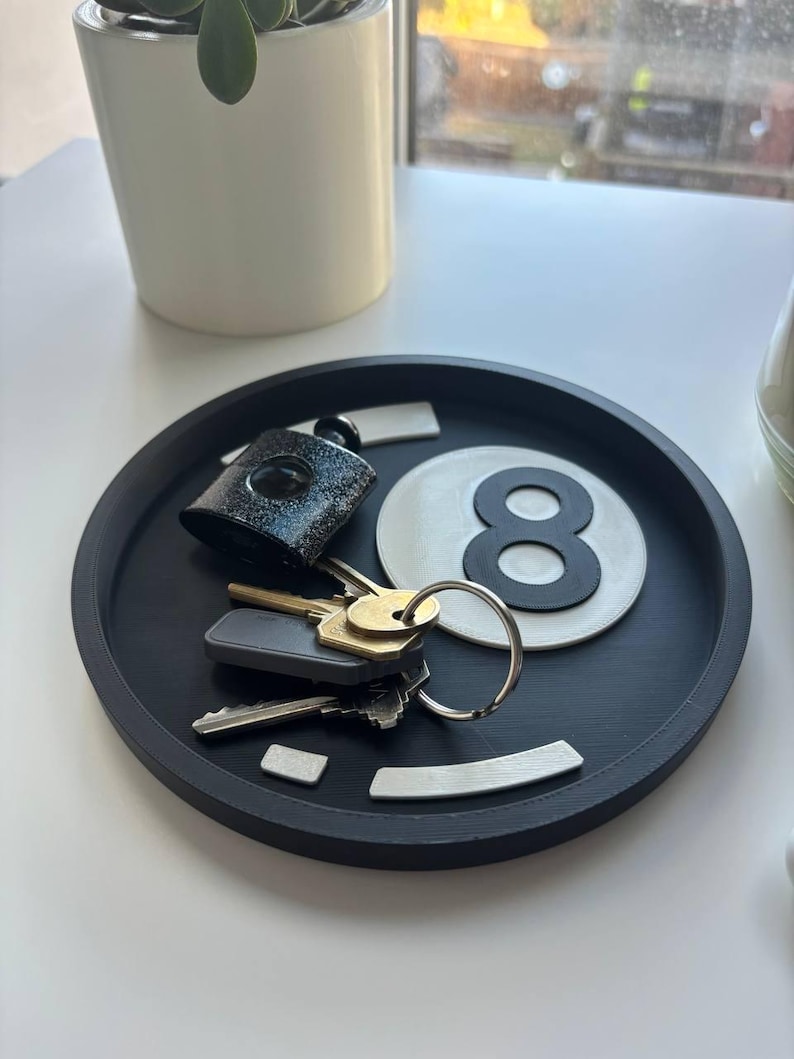 May include: A black circular tray containing keys, a black and silver lock, and a white and black number eight ball. The tray sits on a white surface, with a white plant pot in the background.