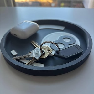 May include: A dark grey round tray containing keys, a white wireless earbud case, and a black tag. The tray has a white and grey design in the centre. The keys are on a silver key ring.