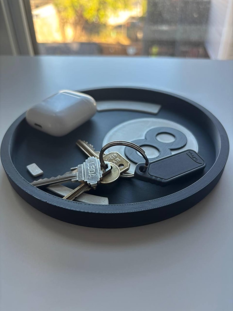 May include: A dark grey round tray containing keys, a white wireless earbud case, and a black tag. The tray has a white and grey design in the centre. The keys are on a silver key ring.