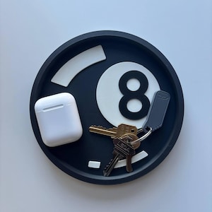 May include: A black and white eight ball-shaped tray, with a pair of white wireless earbuds and a set of keys. The tray has a circular design with a raised edge. The number eight is clearly visible.