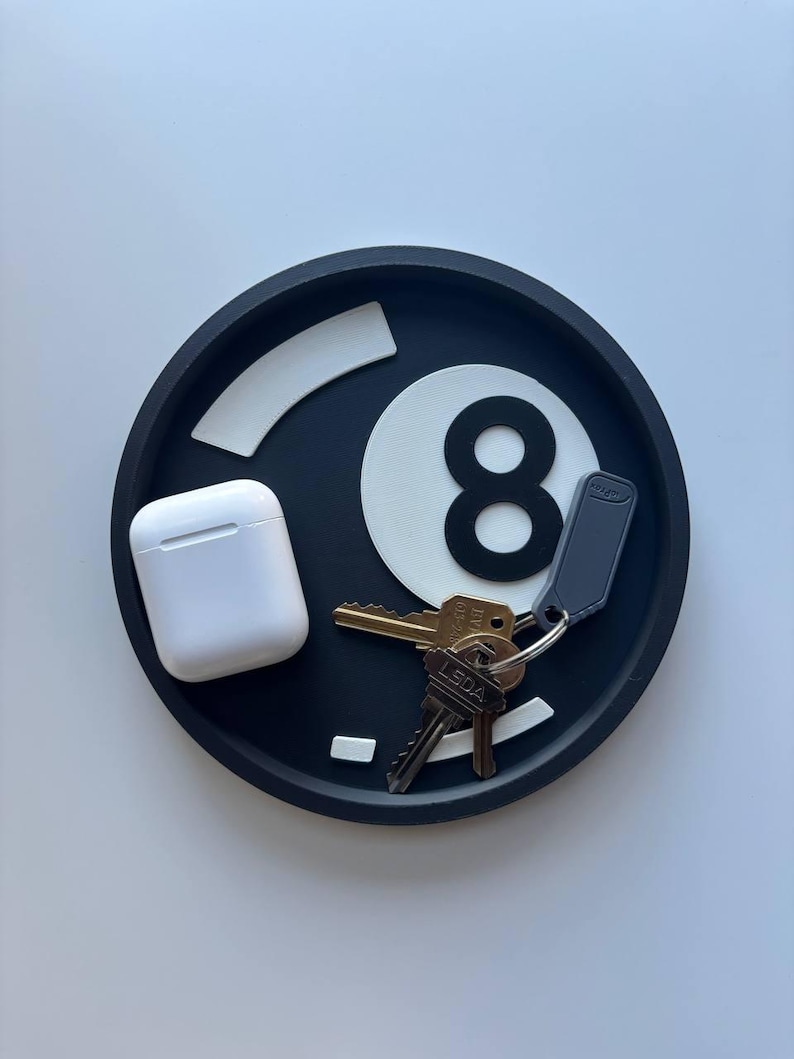 May include: A black and white eight ball-shaped tray, with a pair of white wireless earbuds and a set of keys. The tray has a circular design with a raised edge. The number eight is clearly visible.