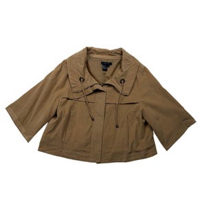 May include: A tan cropped jacket featuring a wide collar, three-quarter length sleeves, and a front placket with a hidden zipper closure. The collar includes two dark brown grommets with thin matching drawstrings that hang down the front.