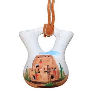May include: A white ceramic pendant with a Native American pueblo design. The pendant has a unique shape with two rounded sections and a central hole. The pueblo is painted in earth tones with green and blue accents, and a brown leather cord.