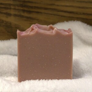 Peut inclure: Un pain de savon artisanal rectangulaire de couleur mauve clair. Le bord supérieur présente une surface texturée et inégale, tandis que les côtés sont lisses. Le savon est posé sur un fond blanc et duveteux.