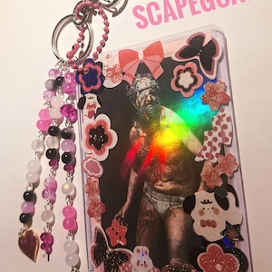 Puede incluir: Un llavero con un tarjetero de pl&aacute;stico transparente con una foto con una figura y la palabra "SCAPEGOAT". El llavero tiene cuentas rosas y negras, un dije en forma de coraz&oacute;n y un cierre plateado en forma de coraz&oacute;n.