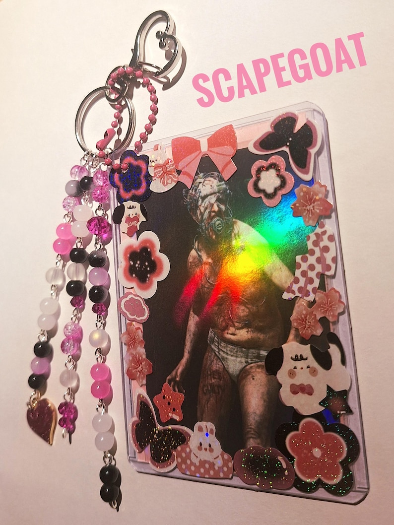 Puede incluir: Un llavero con un tarjetero de pl&aacute;stico transparente con una foto con una figura y la palabra "SCAPEGOAT". El llavero tiene cuentas rosas y negras, un dije en forma de coraz&oacute;n y un cierre plateado en forma de coraz&oacute;n.