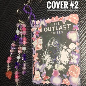 Puede incluir: Un llavero decorativo con una tarjeta con el texto "THE OUTLAST TRIALS". La tarjeta est&aacute; rodeada de pegatinas de mariposas, un conejito y otras formas. El llavero tiene una cadena de cuentas con cuentas rosas, moradas y blancas.