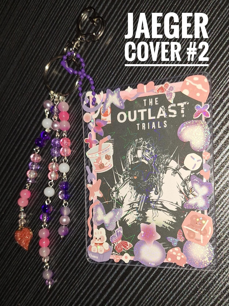 Puede incluir: Un llavero decorativo con una tarjeta con el texto "THE OUTLAST TRIALS". La tarjeta est&aacute; rodeada de pegatinas de mariposas, un conejito y otras formas. El llavero tiene una cadena de cuentas con cuentas rosas, moradas y blancas.