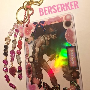 Puede incluir: Un llavero decorativo con una imagen hologr&aacute;fica de un personaje, adornado con detalles en rosa y negro. El llavero incluye un cierre dorado en forma de coraz&oacute;n, hebras de cuentas y la palabra "BERSERKER" en rosa.