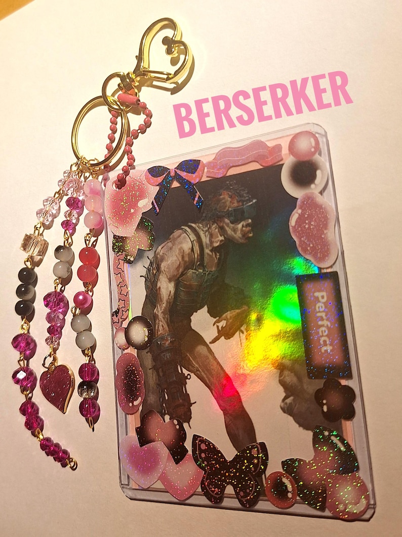 Puede incluir: Un llavero decorativo con una imagen hologr&aacute;fica de un personaje, adornado con detalles en rosa y negro. El llavero incluye un cierre dorado en forma de coraz&oacute;n, hebras de cuentas y la palabra "BERSERKER" en rosa.