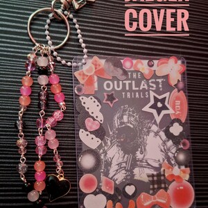 Puede incluir: Un llavero de pl&aacute;stico transparente con un inserto decorativo con el texto "THE OUTLAST TRIALS" y varias pegatinas. El llavero tiene un dije de cuentas con cuentas rosas, naranjas y negras, y un cierre en forma de estrella. Tambi&eacute;n es visible el texto "JAEGER COVER".