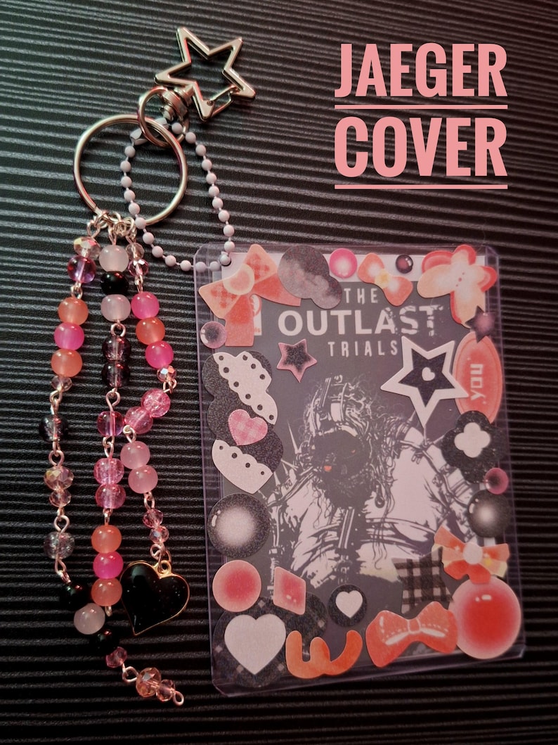 Puede incluir: Un llavero de pl&aacute;stico transparente con un inserto decorativo con el texto "THE OUTLAST TRIALS" y varias pegatinas. El llavero tiene un dije de cuentas con cuentas rosas, naranjas y negras, y un cierre en forma de estrella. Tambi&eacute;n es visible el texto "JAEGER COVER".
