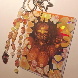 Puede incluir: Un llavero con una tarjeta que muestra una figura enmascarada, adornada con pegatinas de mariposas, lazos y corazones. El llavero incluye un dije de estrella y una cadena de cuentas con un dije de coraz&oacute;n. El texto "PITCHER #3" est&aacute; en la parte inferior.