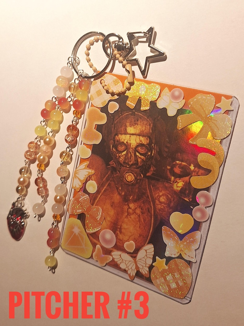 Puede incluir: Un llavero con una tarjeta que muestra una figura enmascarada, adornada con pegatinas de mariposas, lazos y corazones. El llavero incluye un dije de estrella y una cadena de cuentas con un dije de coraz&oacute;n. El texto "PITCHER #3" est&aacute; en la parte inferior.