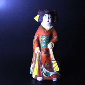 以下が含まれることがあります： 伝統的な日本の着物を着た女性の手描きの陶器人形。人形は主に赤色で、黄色、緑、白のアクセントがあります。扇子を持ち、詳細な髪型をしています。