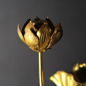 以下が含まれることがあります： 金色の蓮の花の彫刻。花びらのテクスチャが詳細に表現されています。細い金色の茎に取り付けられています。この彫刻はおそらく金属製で、暗い背景に配置されています。