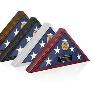 May include: Four triangular flag display cases in brown, black, silver, and cherry wood finishes. Each case holds a folded American flag with a gold United States Army emblem. The cherry case features a black plaque with the text James Anderson.