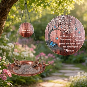 May include: A hanging bird bath with a copper-toned basin and two small bird figures on the rim. Above, a circular plaque reads "In Loving Memory Name Here 1960-2024". A larger circular sign features a tree, blue butterflies, and the text "Your life was a Blessing Your memory a Treasure You are loved beyond words and missed beyond Measure."