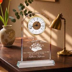 May include: An arched clear acrylic desk clock with a gold-rimmed face featuring Roman numerals. The surface is engraved with the text A NEW CHAPTER BEGINS, a floral book illustration, and the name Katherine Robinson in white script.