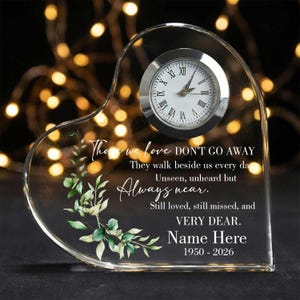 May include: A heart-shaped clear acrylic memorial plaque featuring an inset analog clock with a silver bezel. The surface displays green watercolor foliage and the text: Those we love DON'T GO AWAY They walk beside us every day Unseen, unheard but Always near. Still loved, still missed, and VERY DEAR. Name Here 1950 - 2026.