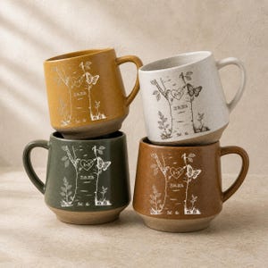 May include: Four ceramic mugs in mustard yellow, speckled white, forest green, and terracotta brown. Each mug features a white line-art design of two birch trees, a heart with the initials A+N, a butterfly, and the date 25.06.2026.