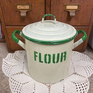 Könnte beinhalten: Ein cremefarbener Mehlbehälter aus Emaille mit passendem Deckel und zwei grünen Griffen. Das Wort FLOUR ist in grünen Blockbuchstaben auf der Vorderseite aufgedruckt. Der Deckel und die Griffe haben grüne Ränder mit sichtbaren Abnutzungsspuren.