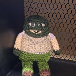 May include: A handmade crochet doll with a green head and body, tan torso, and green pants. The doll has googly eyes, a silver chain necklace, and brown feet. The doll is displayed in a dark corner with a metal mesh background.