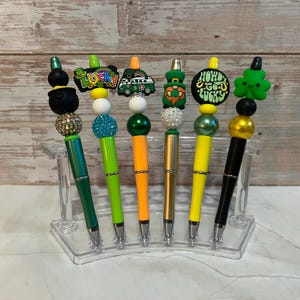 May include: A collection of six decorative pens with St. Patrick's Day themed toppers. Each pen features a different design, including a pot of gold, a four-leaf clover, and the words "Howd Go U Lucky". The pens are displayed in a clear plastic holder.