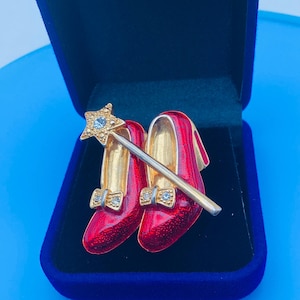 Peut inclure: Broche en forme de chaussures rouges émaillées avec des intérieurs dorés et des détails en forme de nœuds, accompagnée d'une baguette dorée surmontée d'une étoile. La broche est présentée dans un écrin en velours bleu.