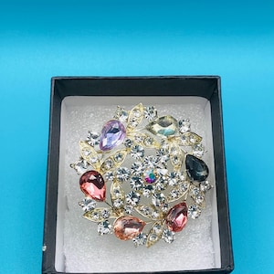 May include: A gold-toned brooch with a floral design, set with clear and colorful gemstones. The brooch features a central cluster of clear crystals surrounded by teardrop-shaped gems in various colors, including pink, purple, and green. It is presented in a black box.