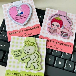 以下が含まれることがあります： 白とパステルカラーのチェック柄台紙にセットされた3つのマグネット式ブックマーク。1つはウサギとテキストが描かれたピンクのハート型、1つはイチゴの頭をしたキャラクターが読書するデザイン、もう1つは猫を乗せた緑色のキャラクターとテキストが描かれています。
