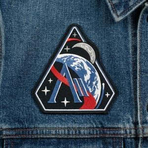 May include: Close-up of a blue denim jacket with a triangular patch. The patch features a stylized Earth, moon, and stars, with the letters "A" and "M" in a red and white design. The jacket has a classic collar and button closures.