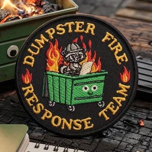 May include: Round black embroidered patch with the words "Dumpster Fire Response Team" in gold lettering. The patch features a raccoon in a dumpster engulfed in flames. The dumpster has googly eyes and is green.
