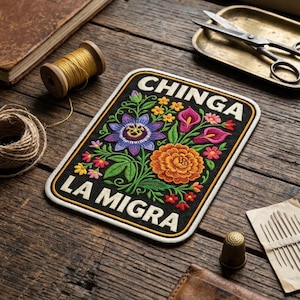 May include: Embroidered patch with the words "CHINGA LA MIGRA" in white, featuring a floral design with a purple passion flower, orange marigold, and pink calla lilies. The patch is rectangular with a black background and a cream border.