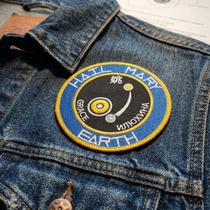 May include: A round embroidered patch with the words "HAIL MARY EARTH" in blue and white, and other text in a foreign language. The patch has a black center with a yellow and white design. The patch is sewn onto a denim jacket.
