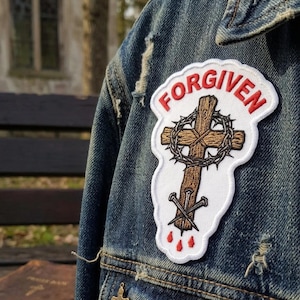 May include: A denim jacket with a patch that reads "FORGIVEN" in red lettering. The patch features a wooden cross with a crown of thorns, nails, and red blood drops. The jacket has a distressed look.