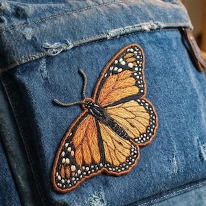 May include: Close-up of a denim jacket with a large, embroidered butterfly patch. The butterfly features orange, black, and white details, with intricate stitching. The jacket is a faded blue color, with distressed details.