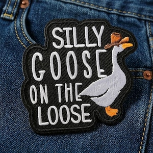 May include: A black embroidered patch with the words "SILLY GOOSE ON THE LOOSE" in white. A cartoon goose wearing a brown cowboy hat is on the right side of the patch. The patch is on a blue denim background.