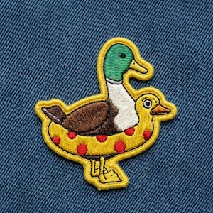 May include: Embroidered patch featuring a duck with a green head, brown body, and white chest, riding a yellow inner tube with red polka dots. The patch has a yellow border and is set against a blue denim background.