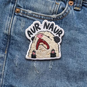 May include: Embroidered patch featuring a distressed hamster with the text "AUR NAVR" in a circular design. The hamster has a pink nose, open red mouth, and black paws and ears. The patch is on a blue denim background.