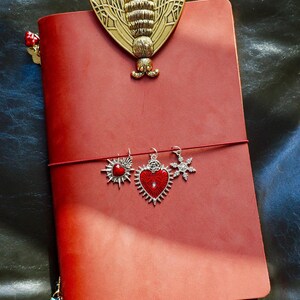 May include: A red leather journal with a gold-colored moth-shaped clip and three charms: a sunburst with a red center, a heart with a red center, and a snowflake. The journal is closed with a red elastic band.