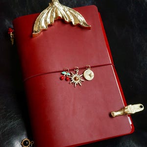 May include: A red leather journal with gold-colored mermaid tail and charm accents. The journal features a gold sun, star, and cherry charm. A gold alligator clip and a small black and blue charm are also visible.
