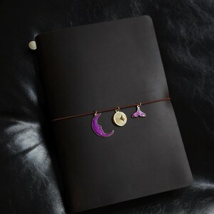 May include: A dark brown leather journal with a brown elastic closure. The journal is adorned with three decorative charms: a purple crescent moon, a gold compass, and a purple mermaid tail. The journal is closed.