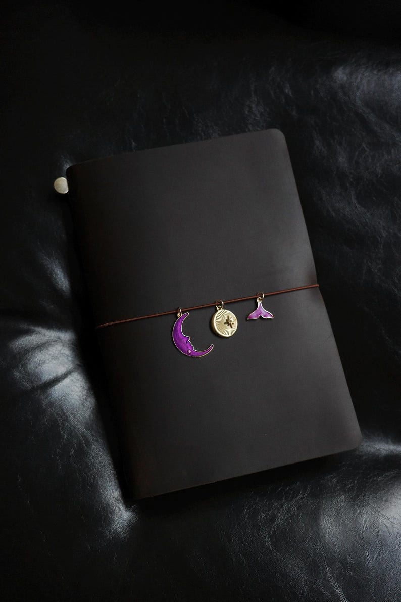May include: A dark brown leather journal with a brown elastic closure. The journal is adorned with three decorative charms: a purple crescent moon, a gold compass, and a purple mermaid tail. The journal is closed.